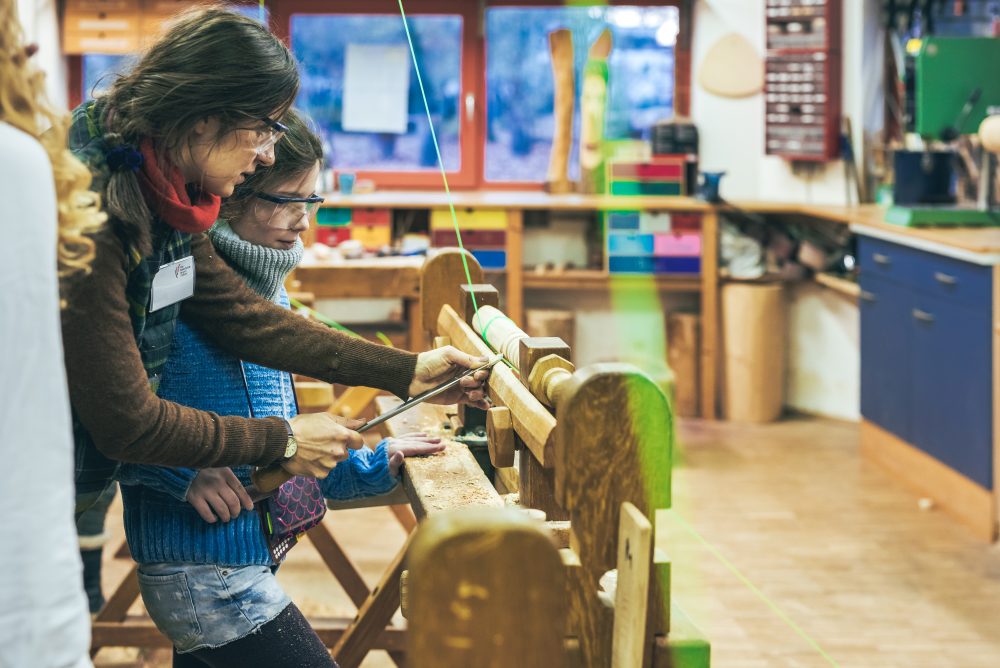 Zwei Frauen arbeiten in einer Holzwerkstatt und benutzen Werkzeuge, um ein Holzprojekt zu bearbeiten. Die Atmosphäre ist kreativ und lehrreich, während sie gemeinsam handwerkliche Fähigkeiten erlernen.
